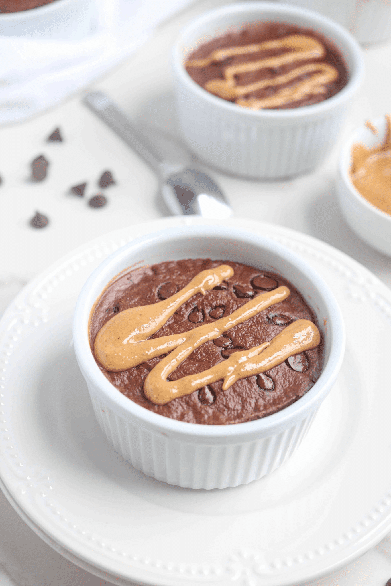 chocolate baked oats in a white ramekin on a white plate with chocolate chips and drizzled with peanut butter.