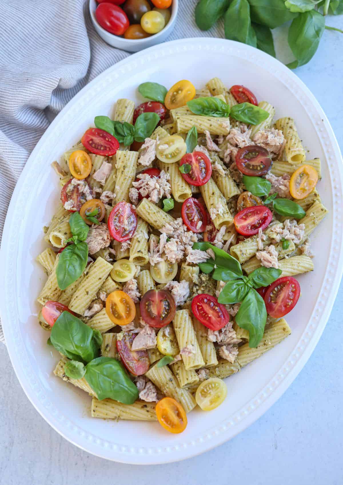 pesto tuna pasta with cherry tomatoes, tuna and fresh basil leaves on an oval white platter.