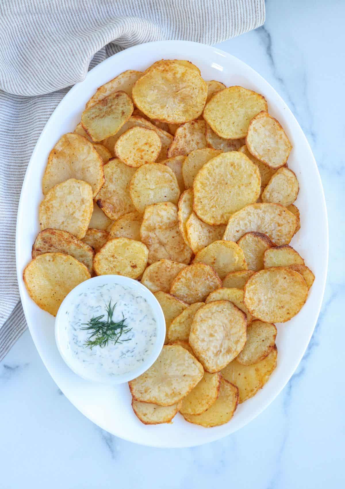 air fried potato slices on a white oval platter with a side of sill aioli in a small round white bowl. 