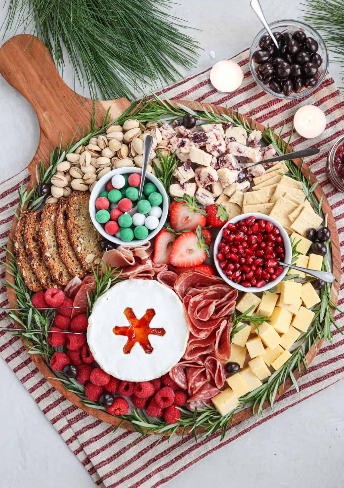 round Christmas charcuterie board featuring 3 kinds of cheese, meats, fruit, nuts and candy with a rosemary rim.