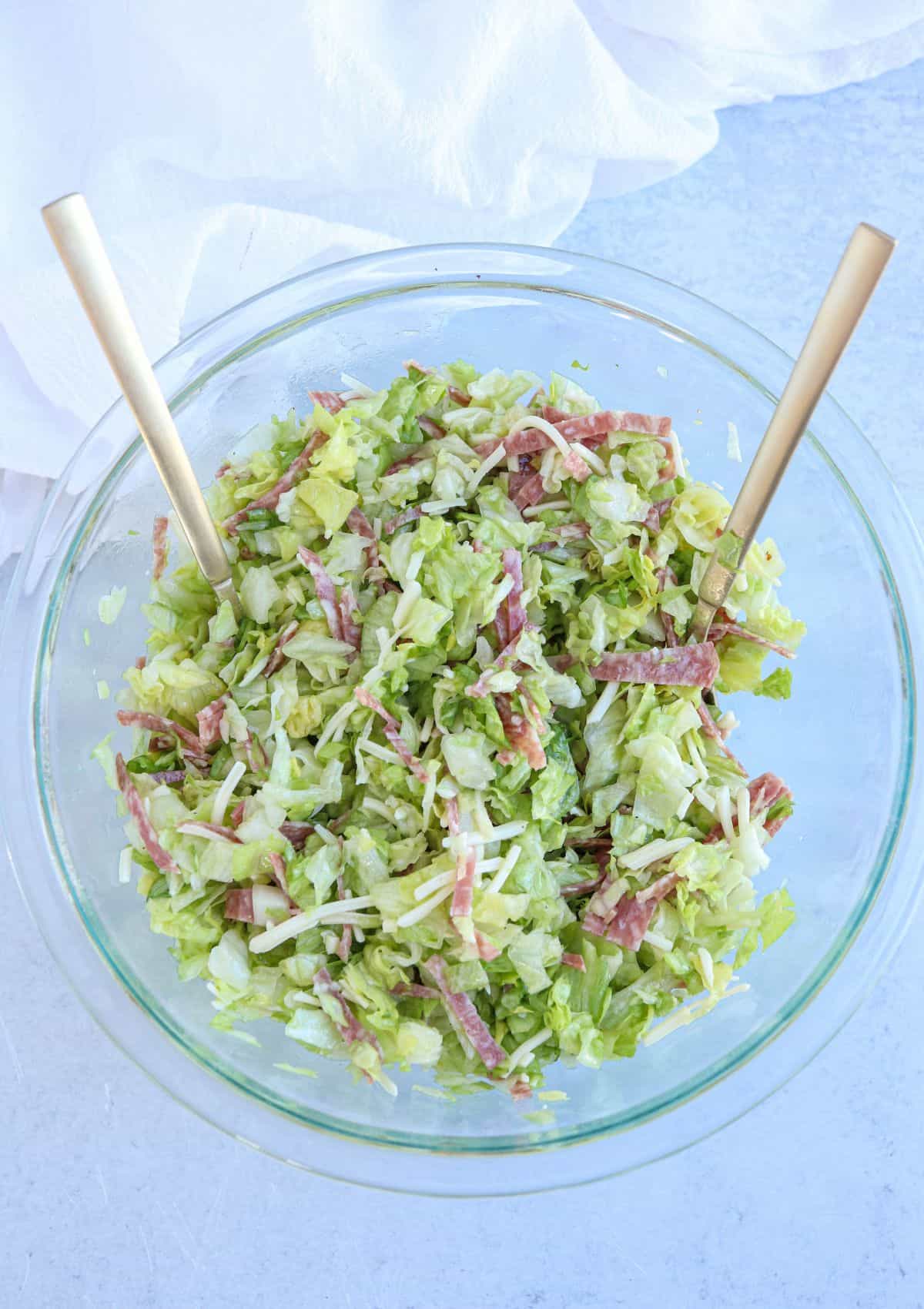 overhead view of a chopped salad with salami, mozzarella in a glass bowl with golden spoons.