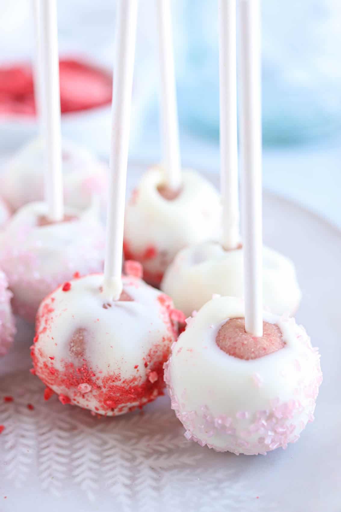 strawberry cake pops coated in white chocolate on a plate, white sticks upward.