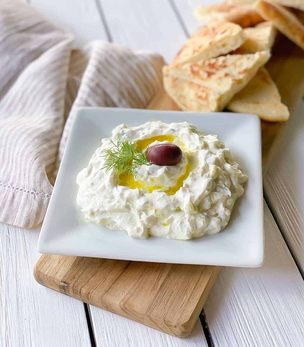 thick tzatziki sauce with olive oil, a single olive and a sprig of fresh dill on a square white plate.