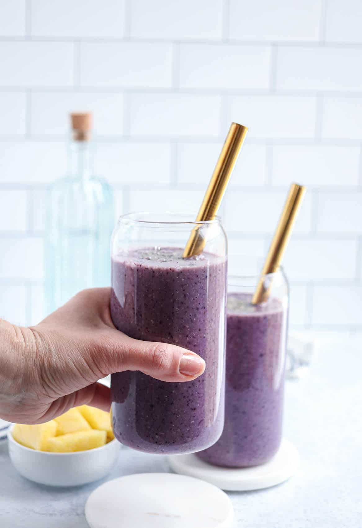 two glasses of blueberry pineapple smoothie with gold straws on light gray surface, one glass being liften by a hand.
