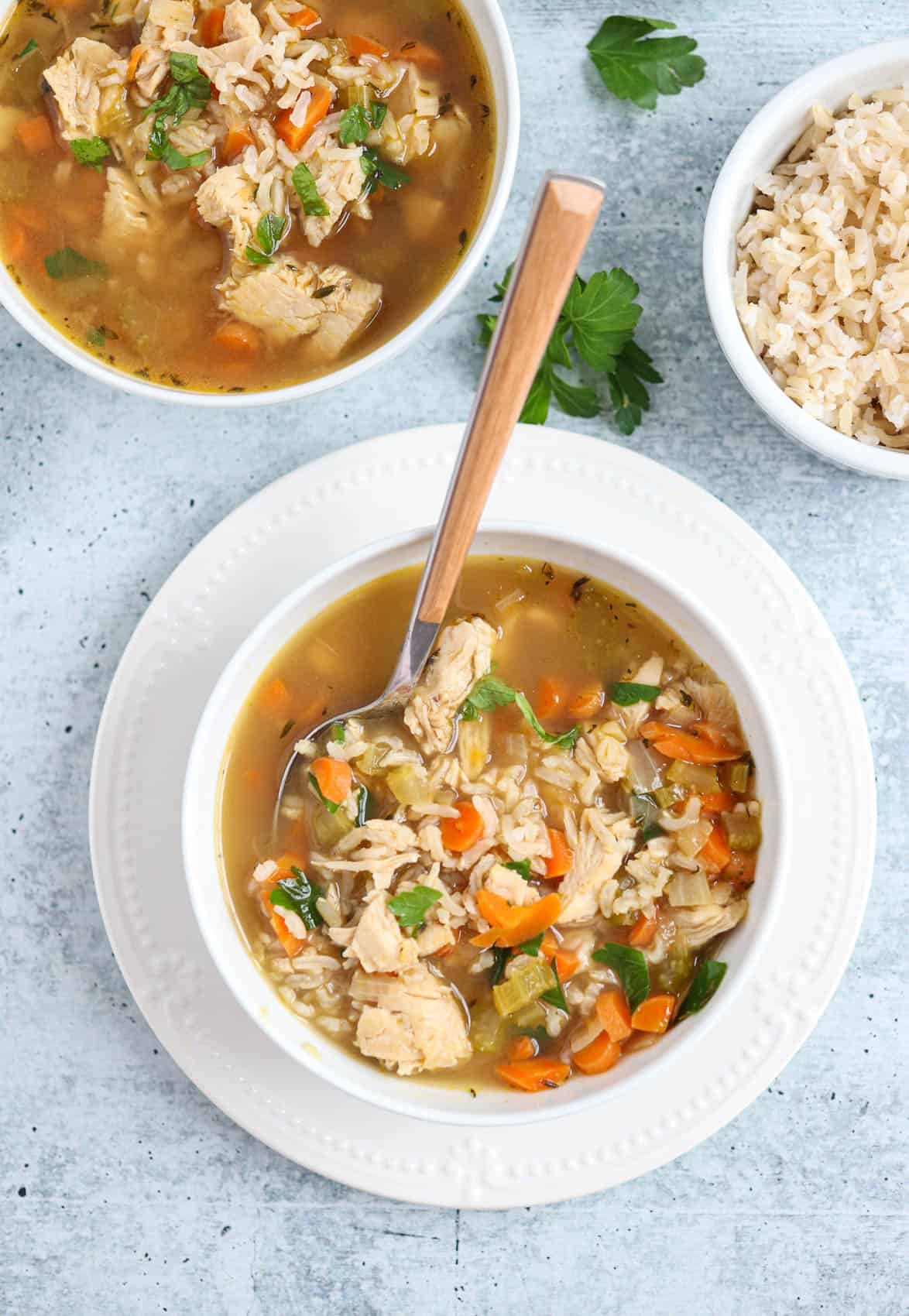 bone broth chicken soup with carrots and brown rice in a white bowl on a white plate with a spoon inserted in the soup.