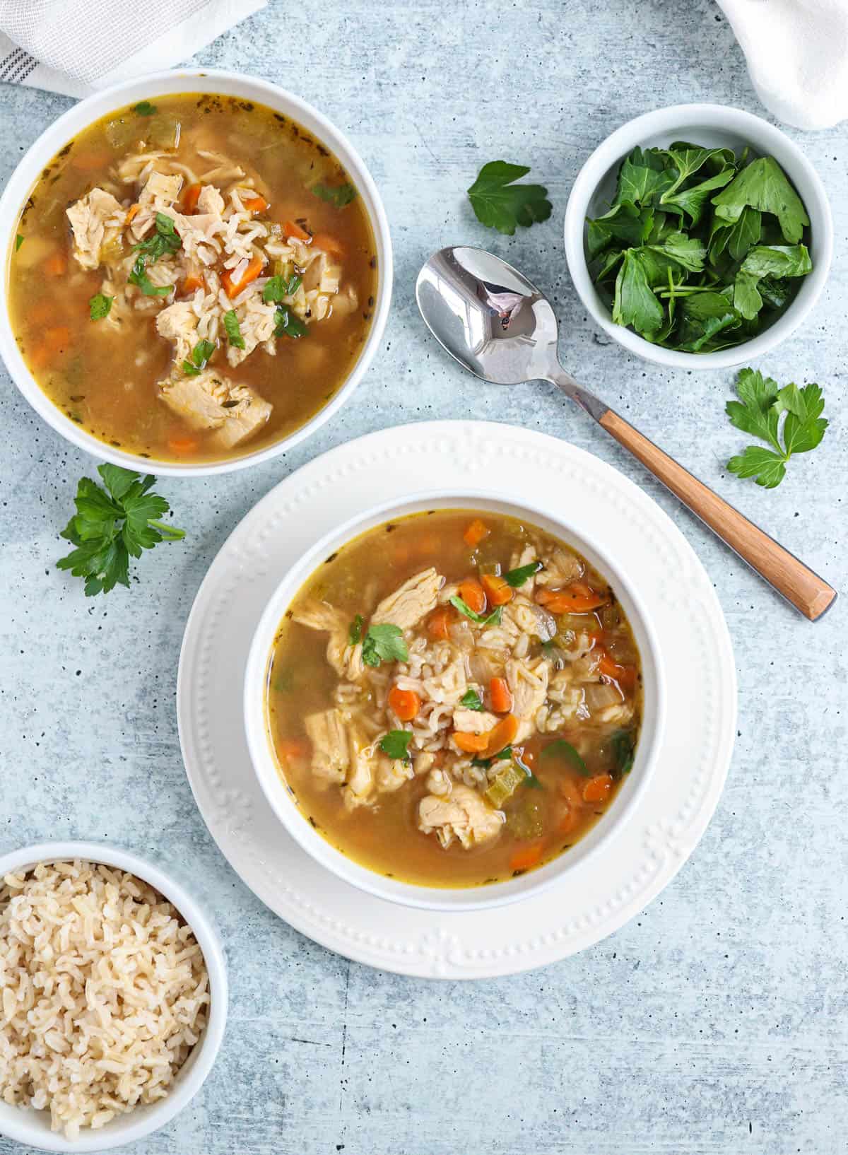 bone broth chicken soup with carrots and brown rice in a white bowl on a white plate on gray surface with another bowl of soup and a spoon nearby.