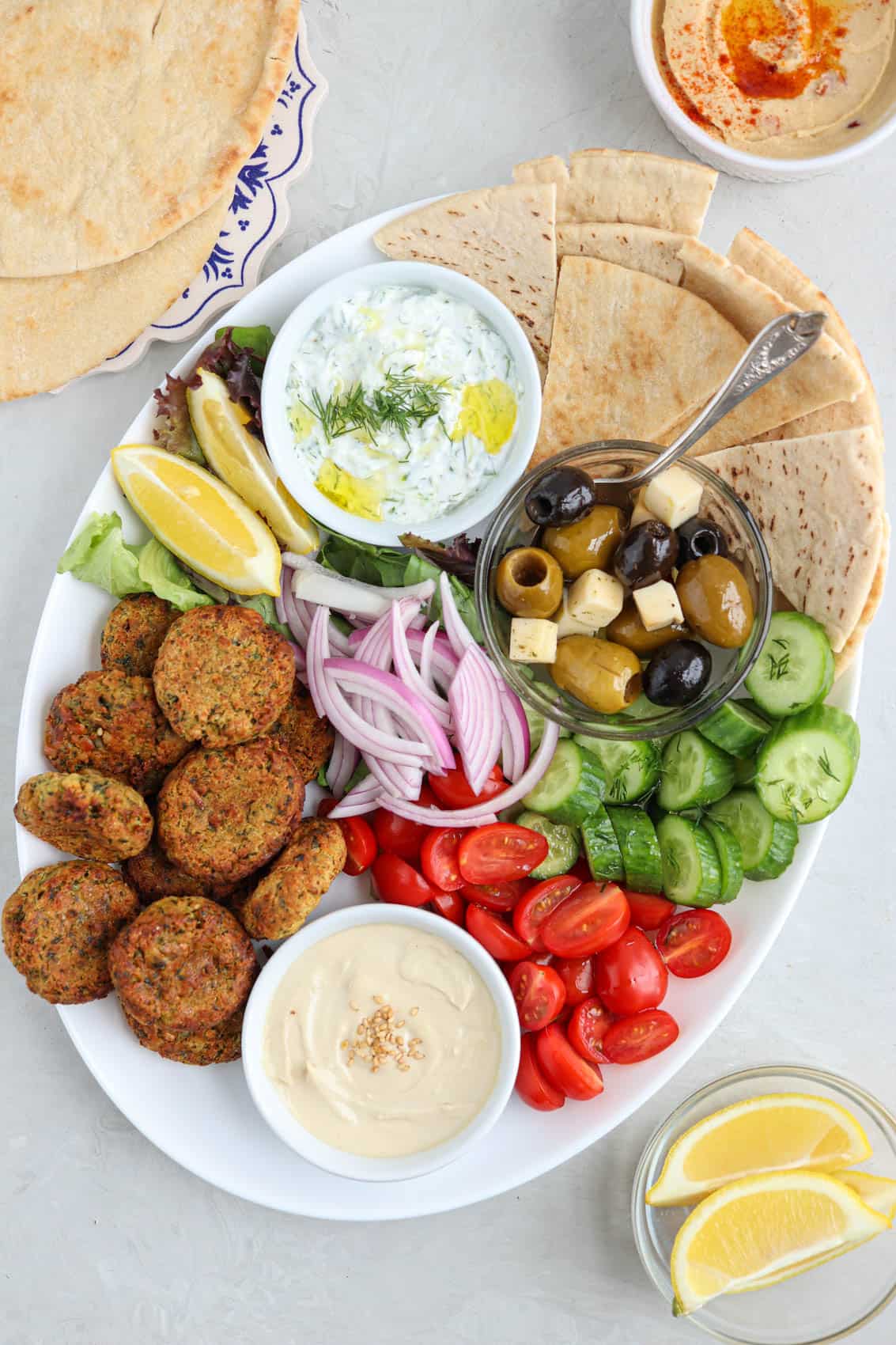 oval falafel platter with falafel patties, pita triangles, fresh veggies, two dips, greens and olives.
