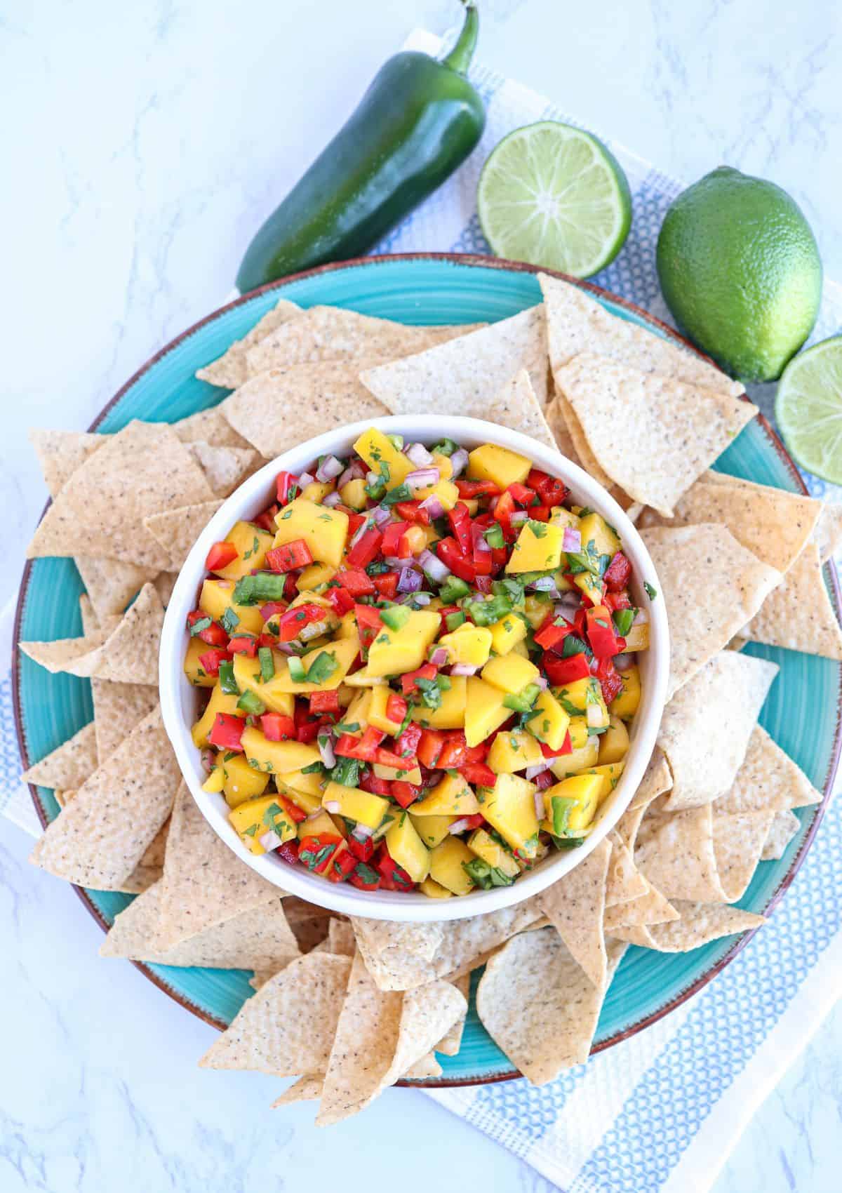 fresh mango pico de gallo in a white and blue bowl surrounded by tortilla chips on a blue plate.