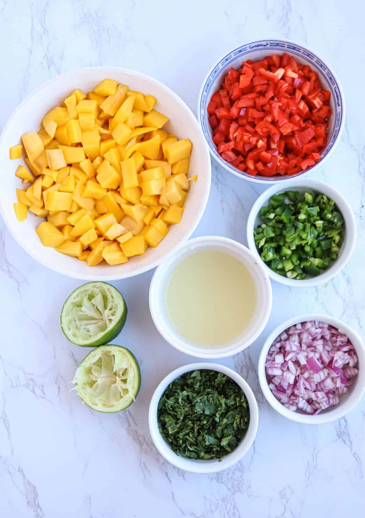 ingredients for making fresh mango salsa in round containers on white marble surface.