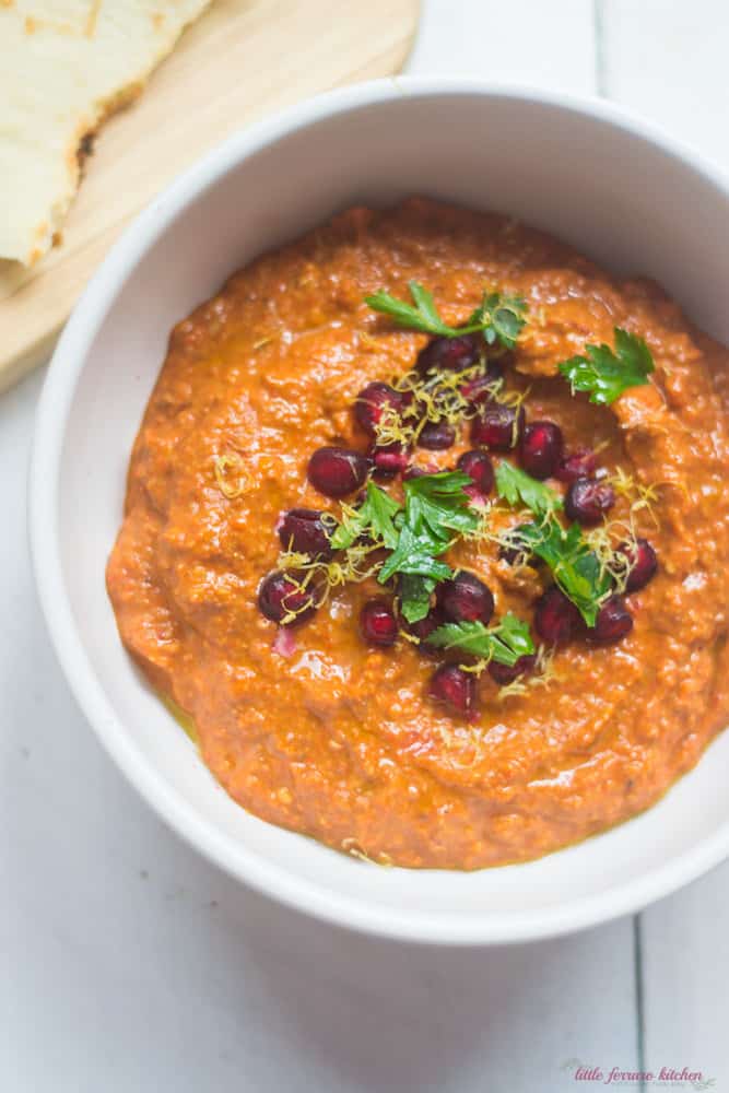 orange thick sauce in a white bowl garnished with pomegranate seeds, parsley and lemon peel.