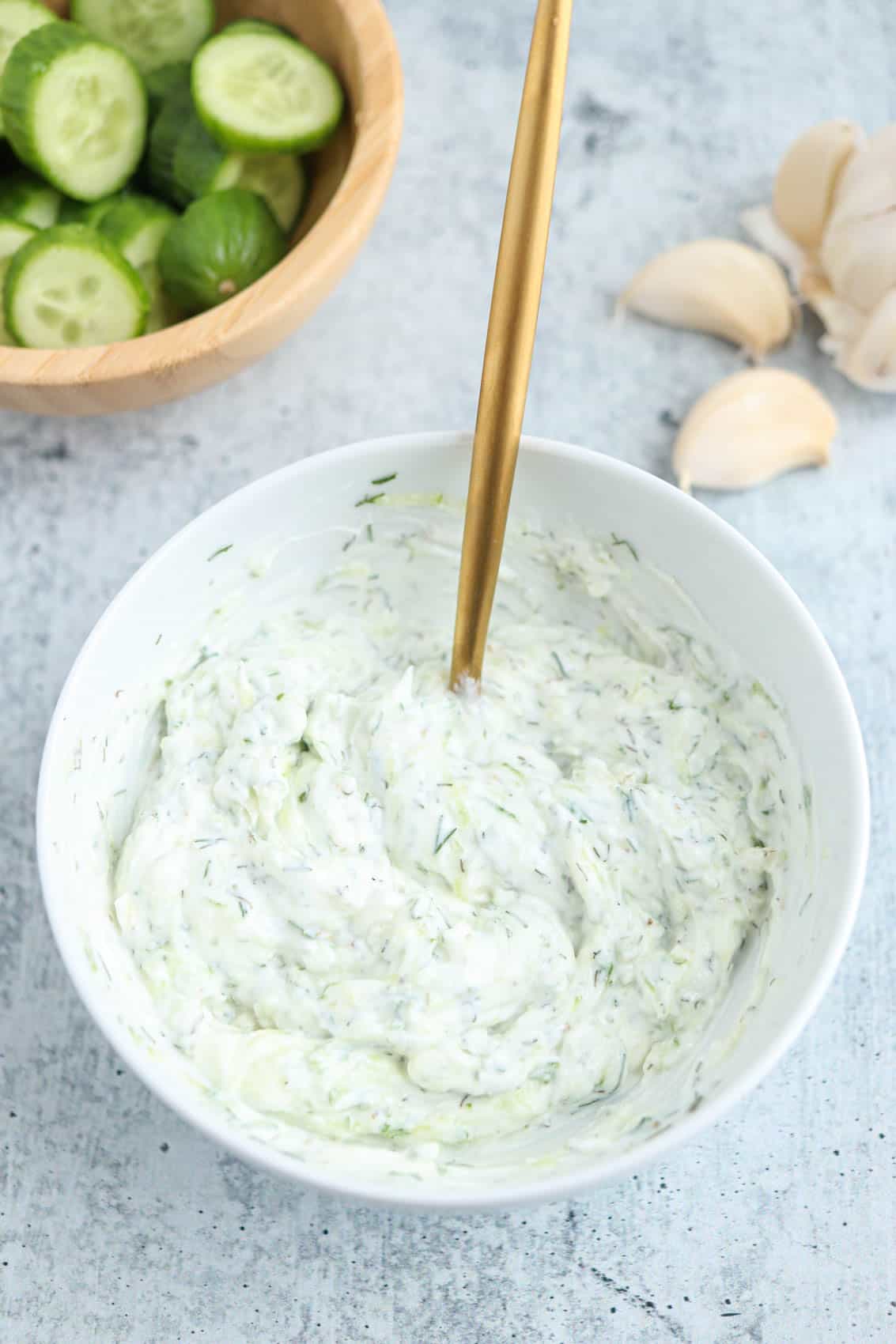 a white bowl with tzatziki yogurt sauce on gray surface with a golden spoon inside the bowl.