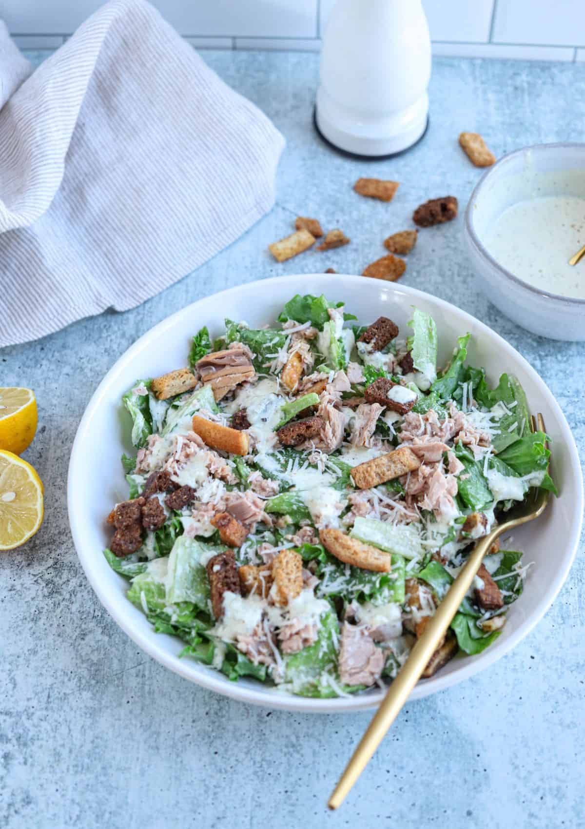 caesar salad wth coutons , tuna, parmesan cheese and dressing in a white bowl.