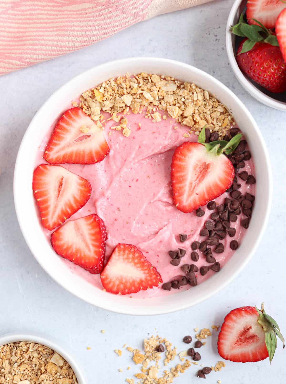 strawberry smoothie bowl in a white bowl topped with fresh strawberries, mini chocolate chips and granola.