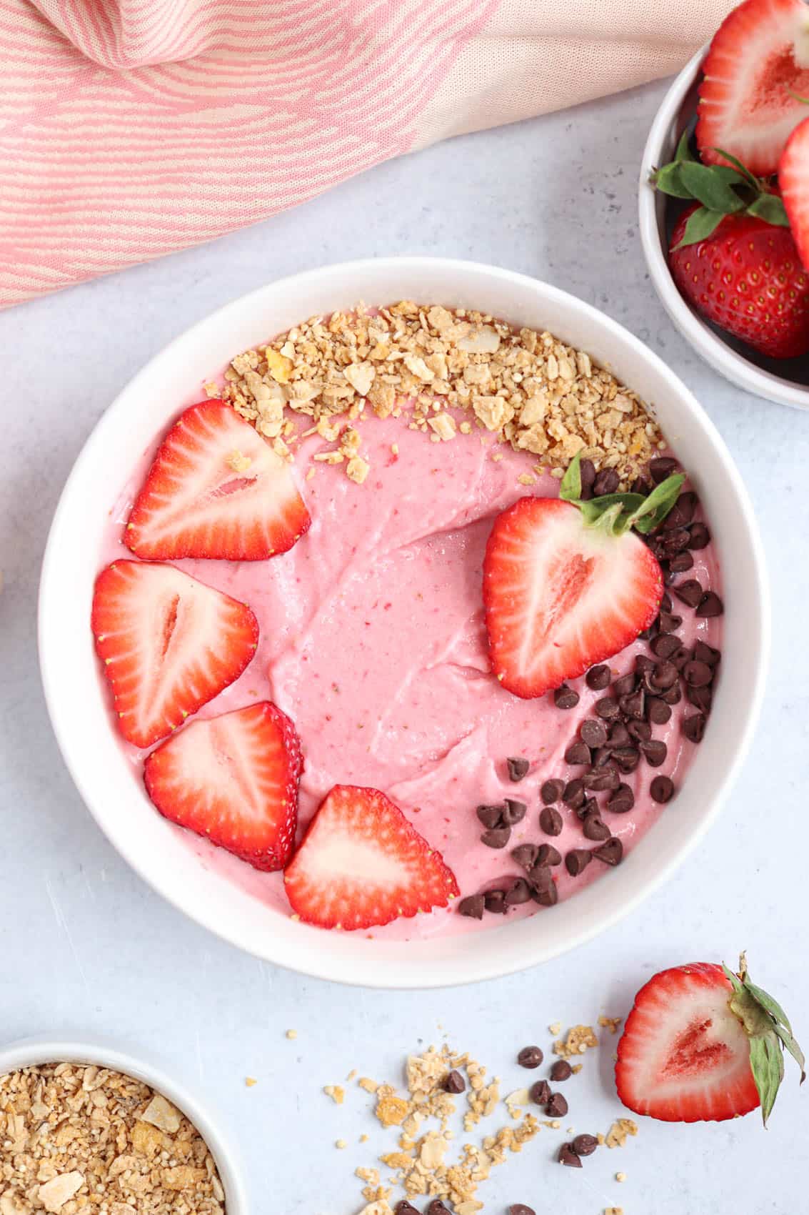 strawberry smoothie bowl in a white bowl topped with fresh strawberries, mini chocolate chips and granola.