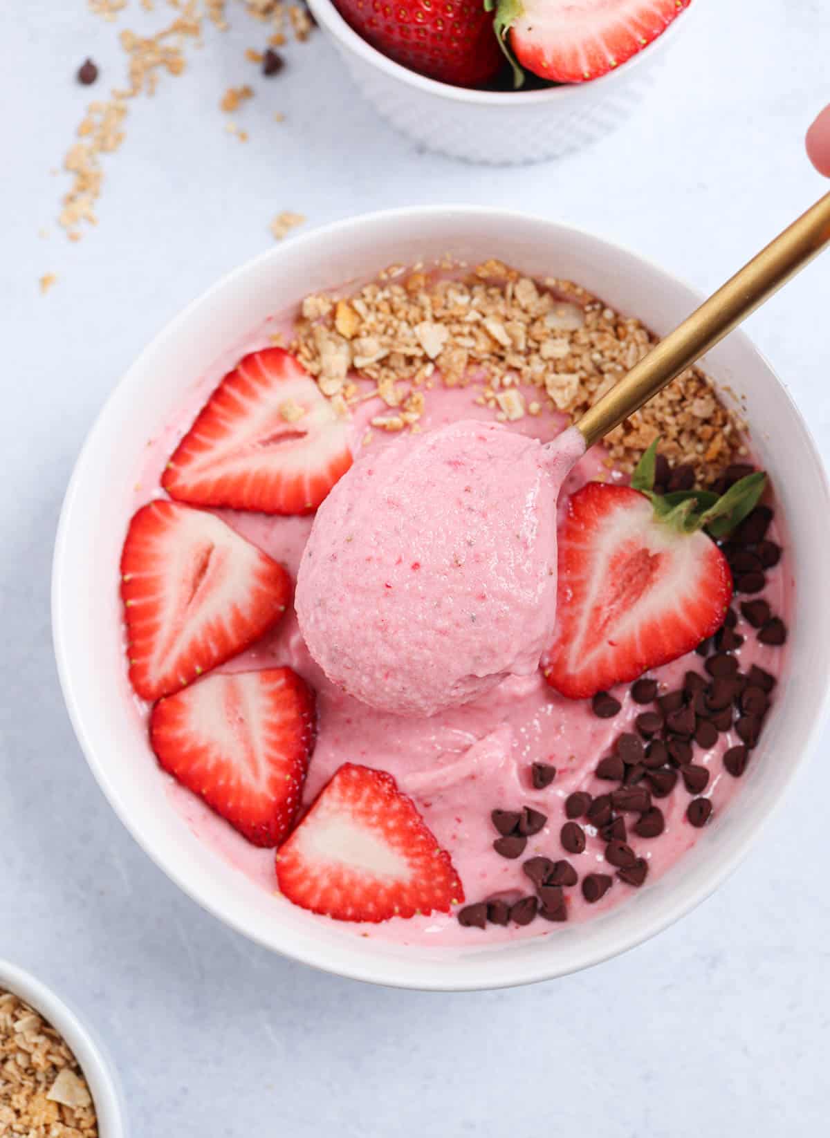 strawberry smoothie bowl in a white bowl topped with fresh strawberries, mini chocolate chips and granola with a spoonful close-up.