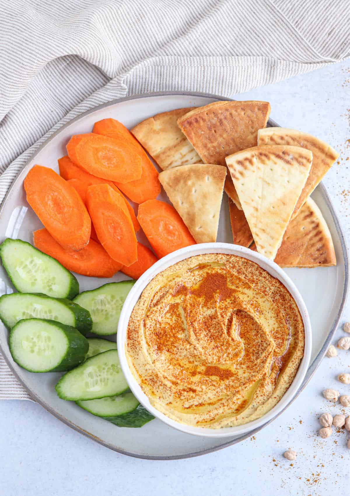 spiced hummus in a bowl topped with smoked paprika and olive oil surrounded by pita chips and fresh veggies.