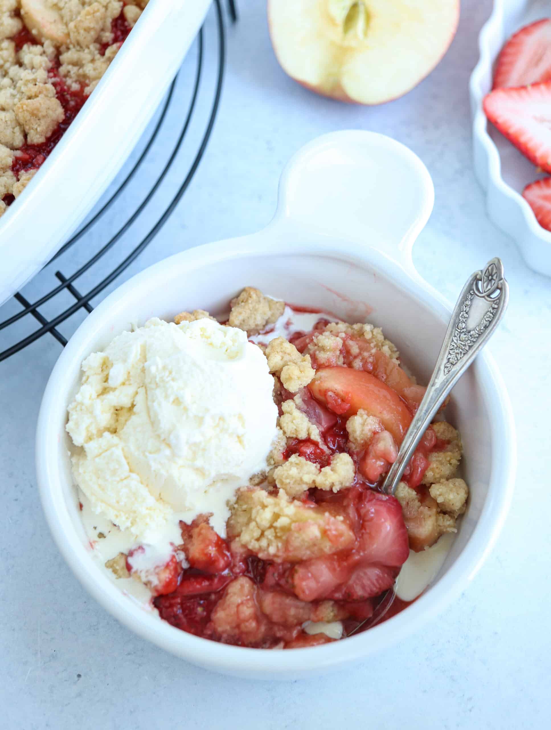 strawberry apple crisp in a white bowl with a scoop of ice cream and a spoon. 