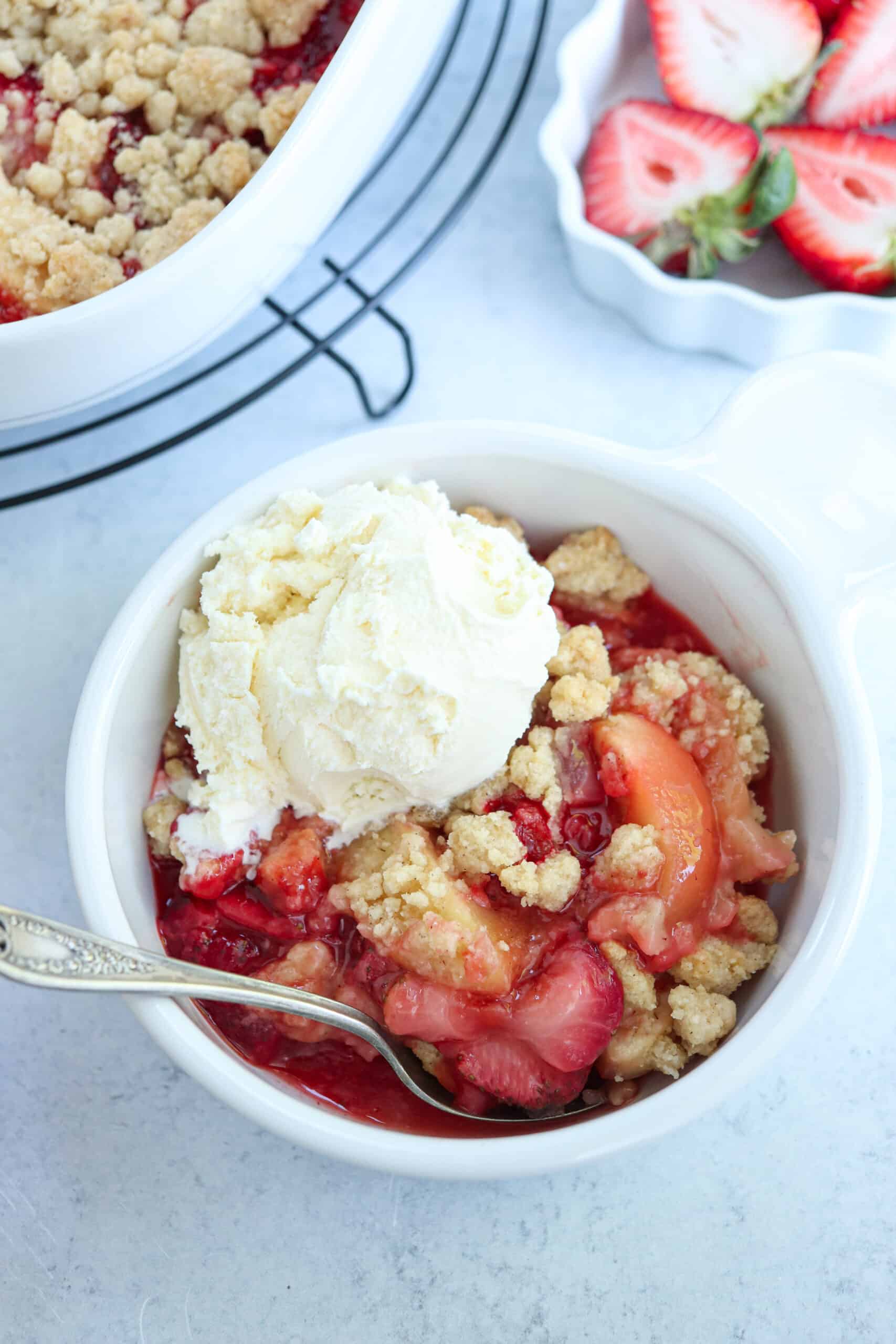 strawberry apple crisp in a white bowl with a scoop of ice cream and a spoon.