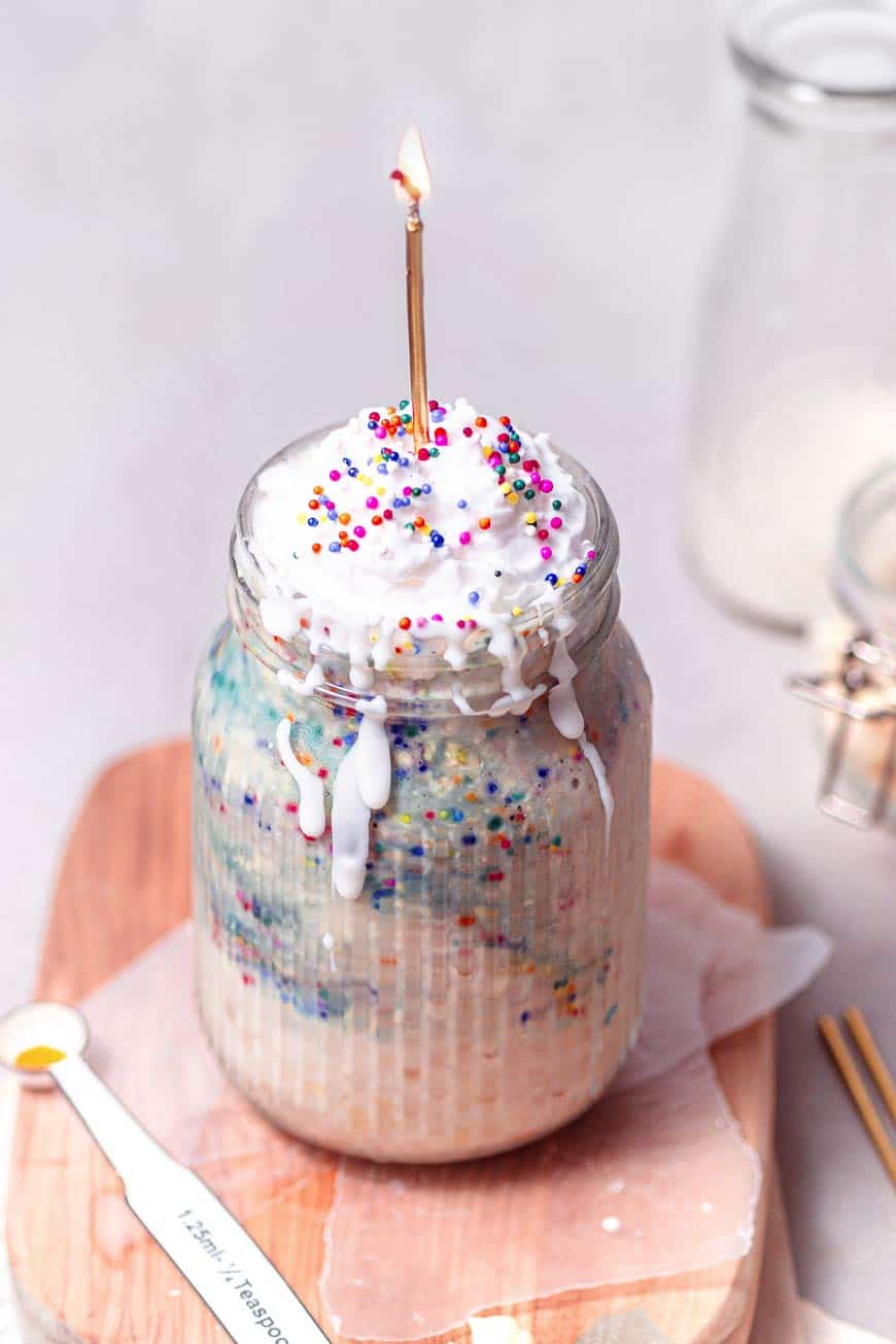 overnight oats in a mason jar topped with whipped cream, rainbow sprinkles and a golden candle. 