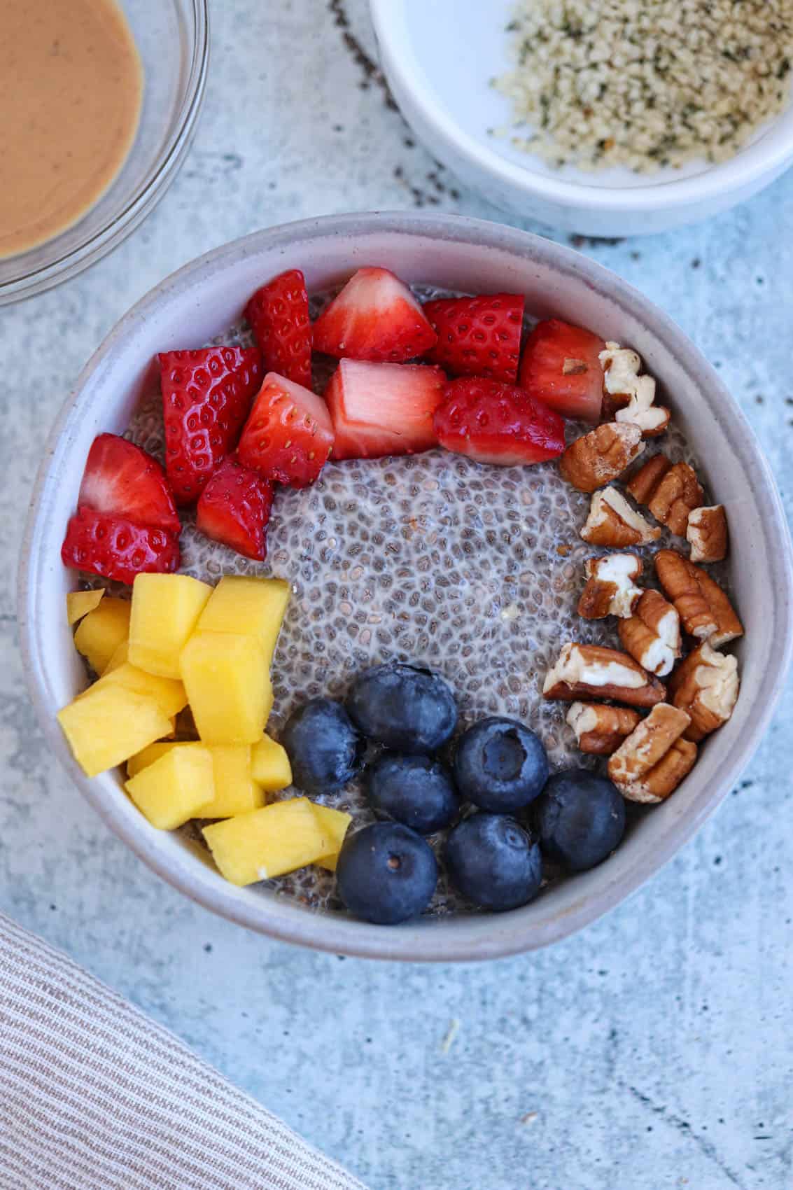 gray bowl with chia pudding, fresh fruit and pecans on a light gray surface. 