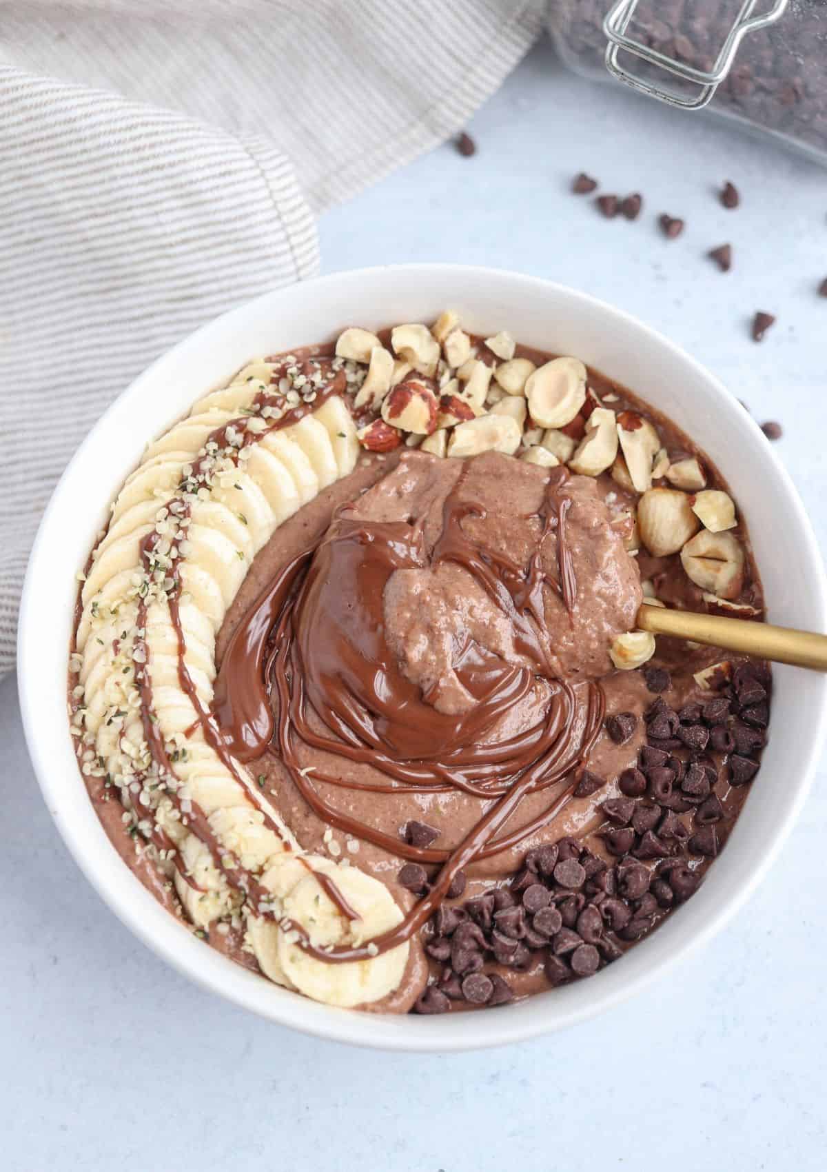 nutella smoothie bowl in a white bowl topped with banana slices, hazelnuts, nutella and chocolate chips. 
