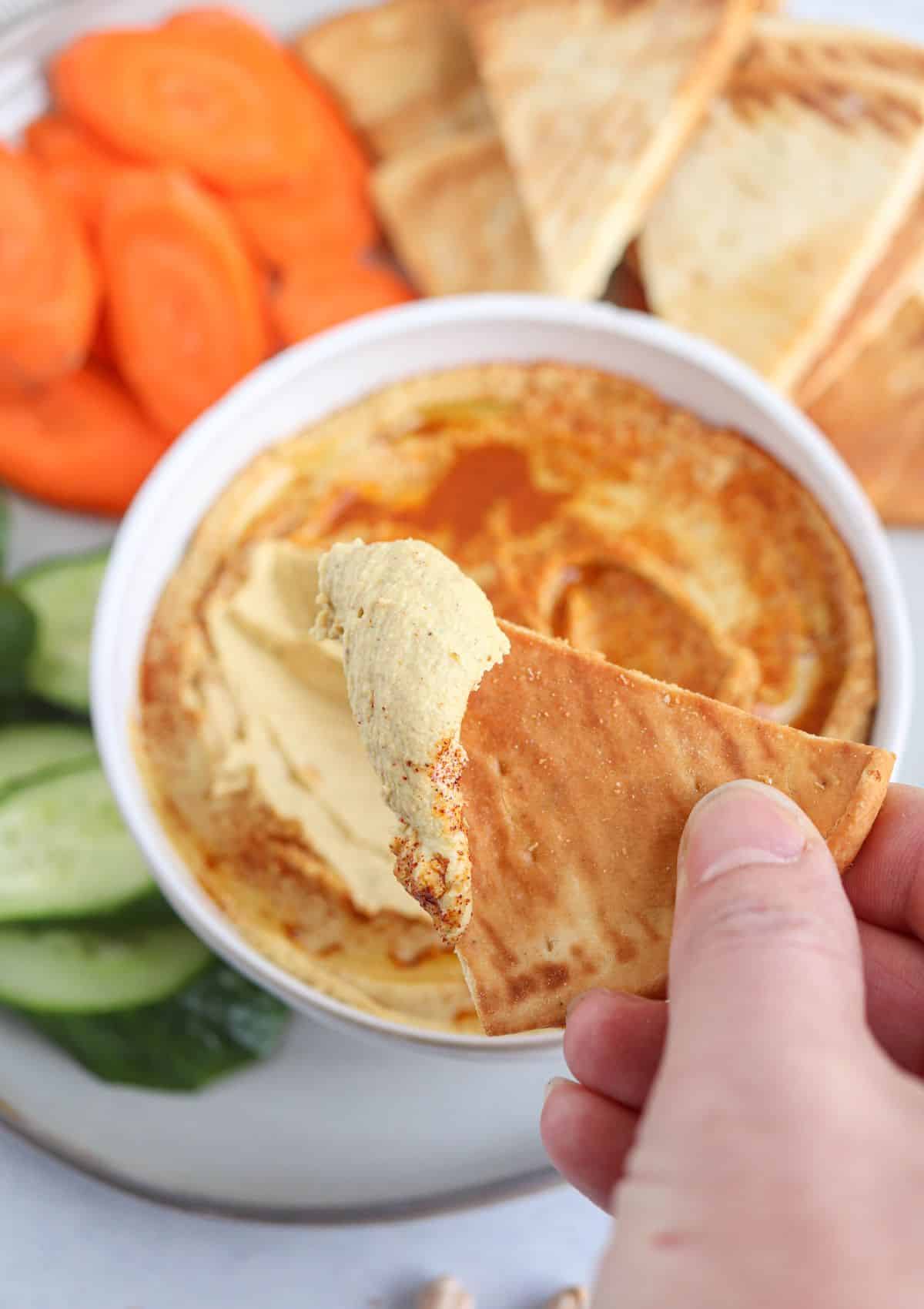 close up of spiced hummus on a pita chip and in a bowl topped with smoked paprika and olive oil surrounded by pita chips and fresh veggies.