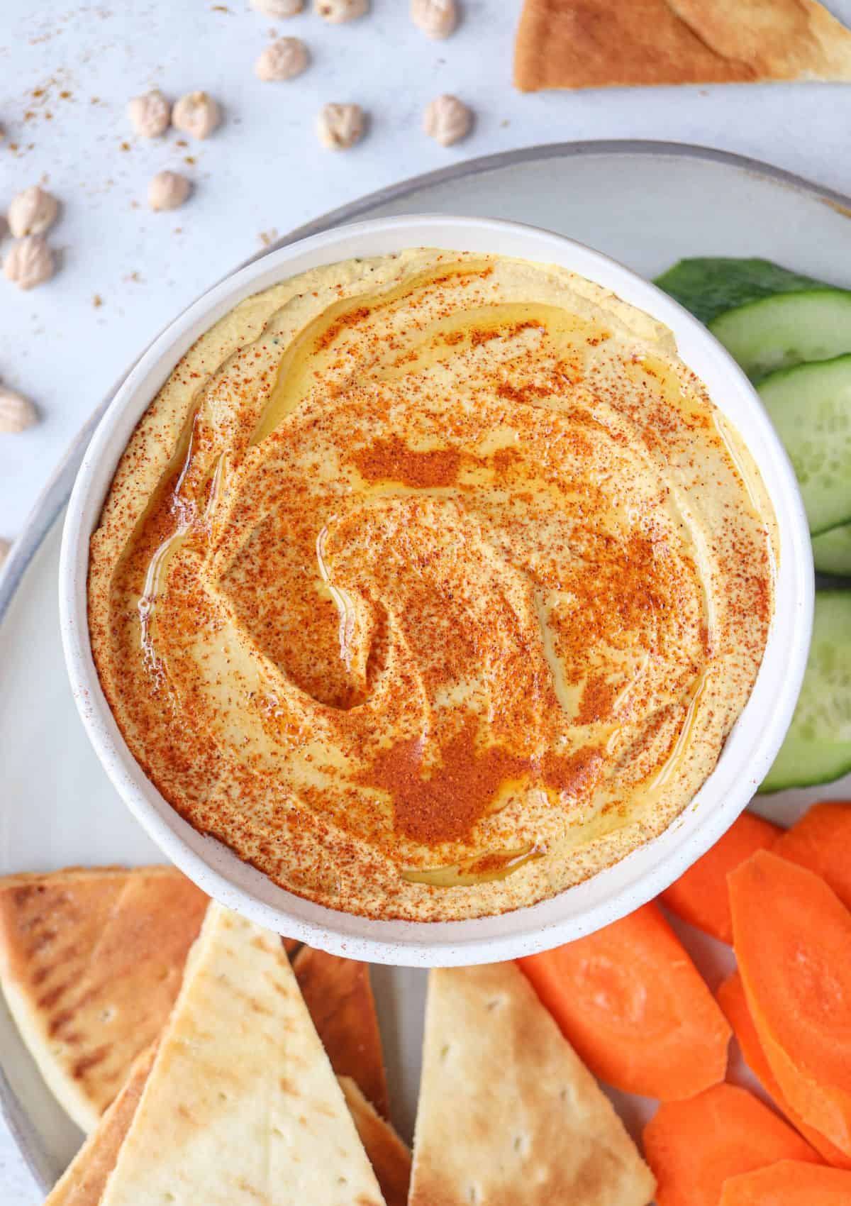 spiced hummus in a bowl topped with smoked paprika and olive oil surrounded by pita chips and fresh veggies.