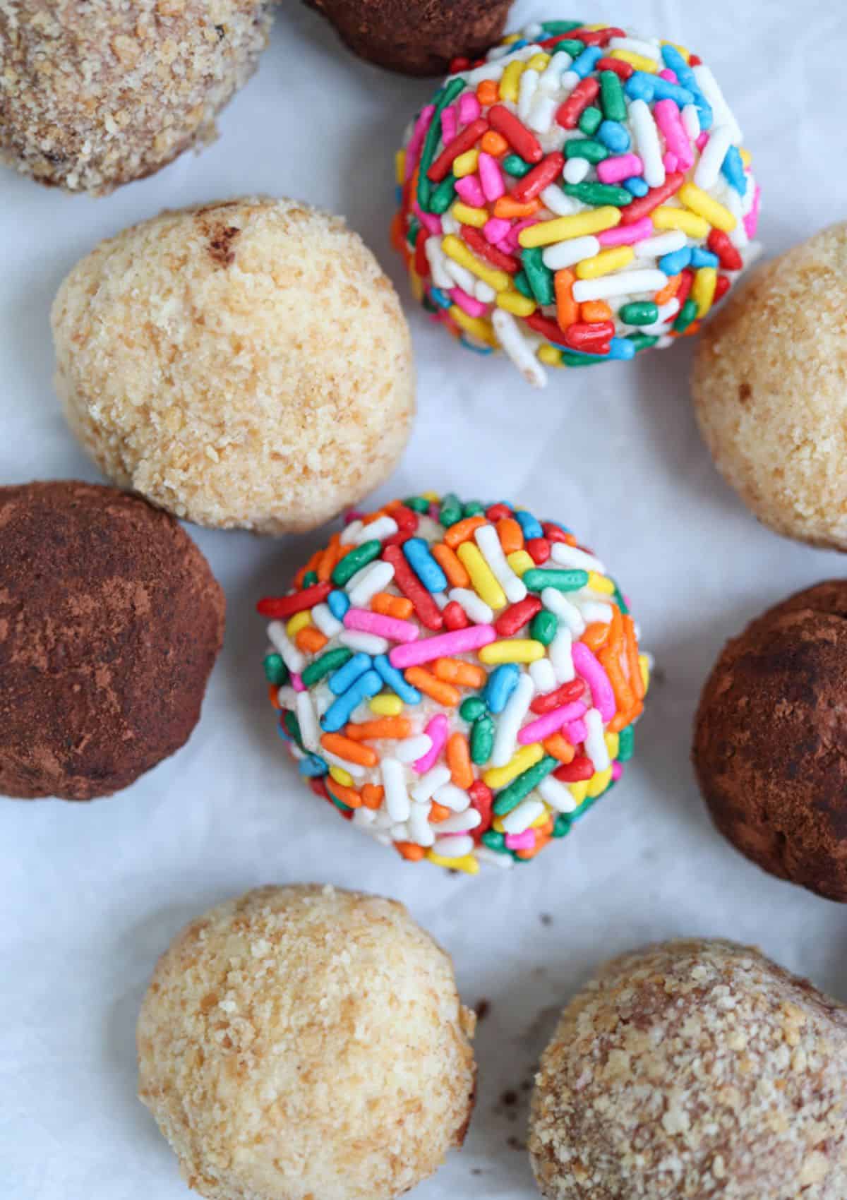 cheesecake balls coated in rainbow sprinkles, cookie crumbs and cocoa powder on parchment paper.