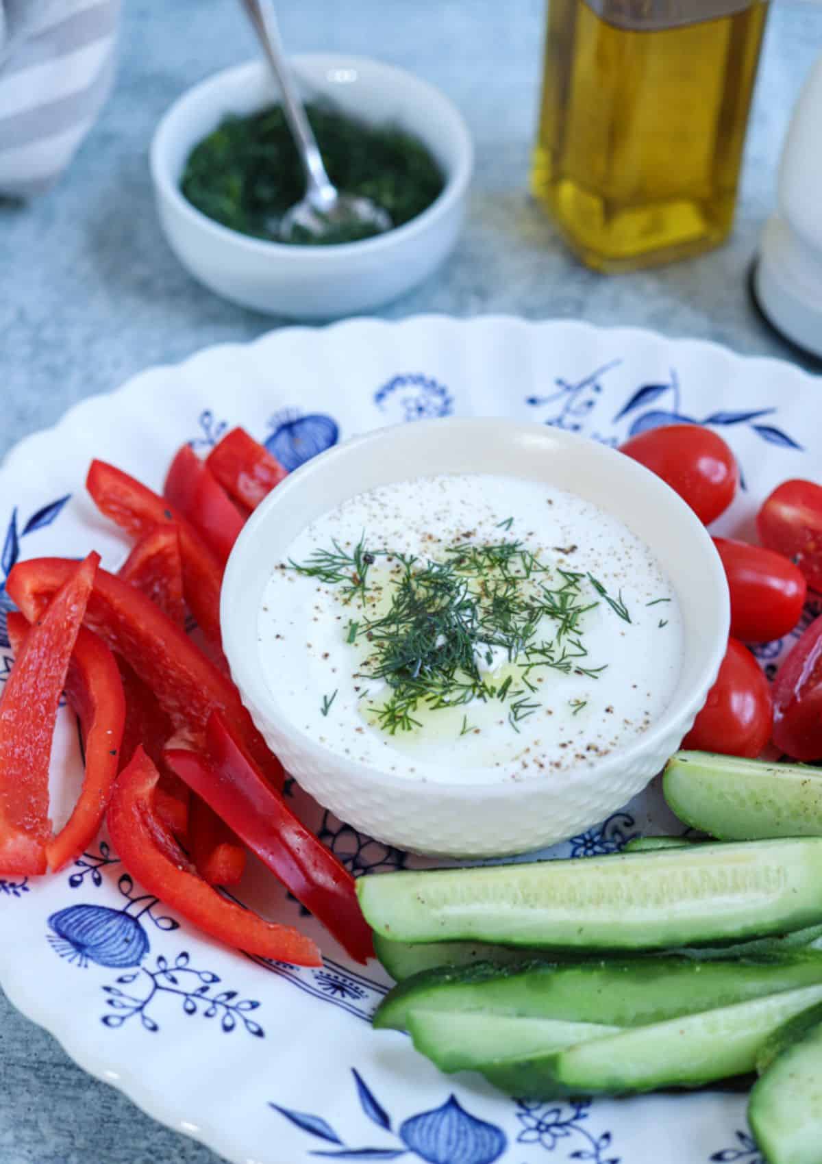 blended cottage cheese sprinkled with fresh dill and black pepper on a small white bowl surrounded by veggies on a platter. 