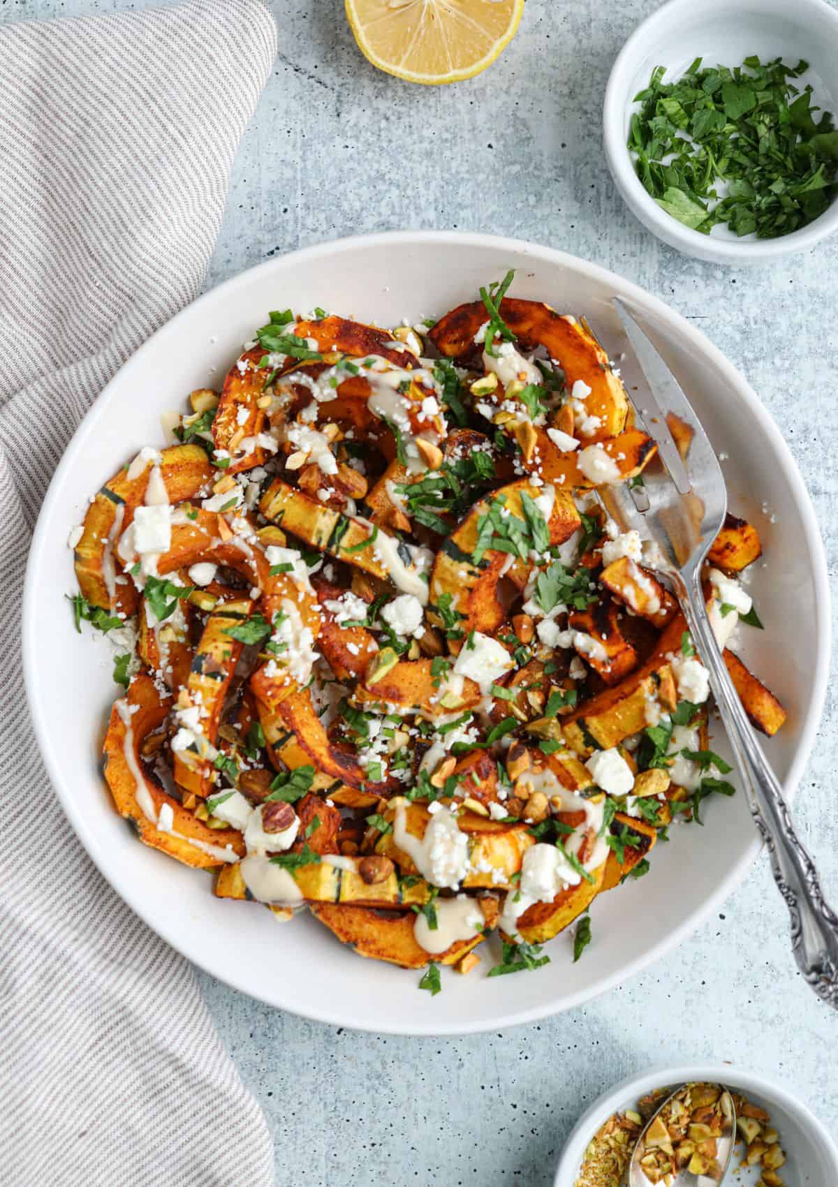 sautéed delicata squash drizzled with lemon tahini dressing and sprinkled with chopped parsley, crumbled feta cheese and pistachios in a white bowl. 