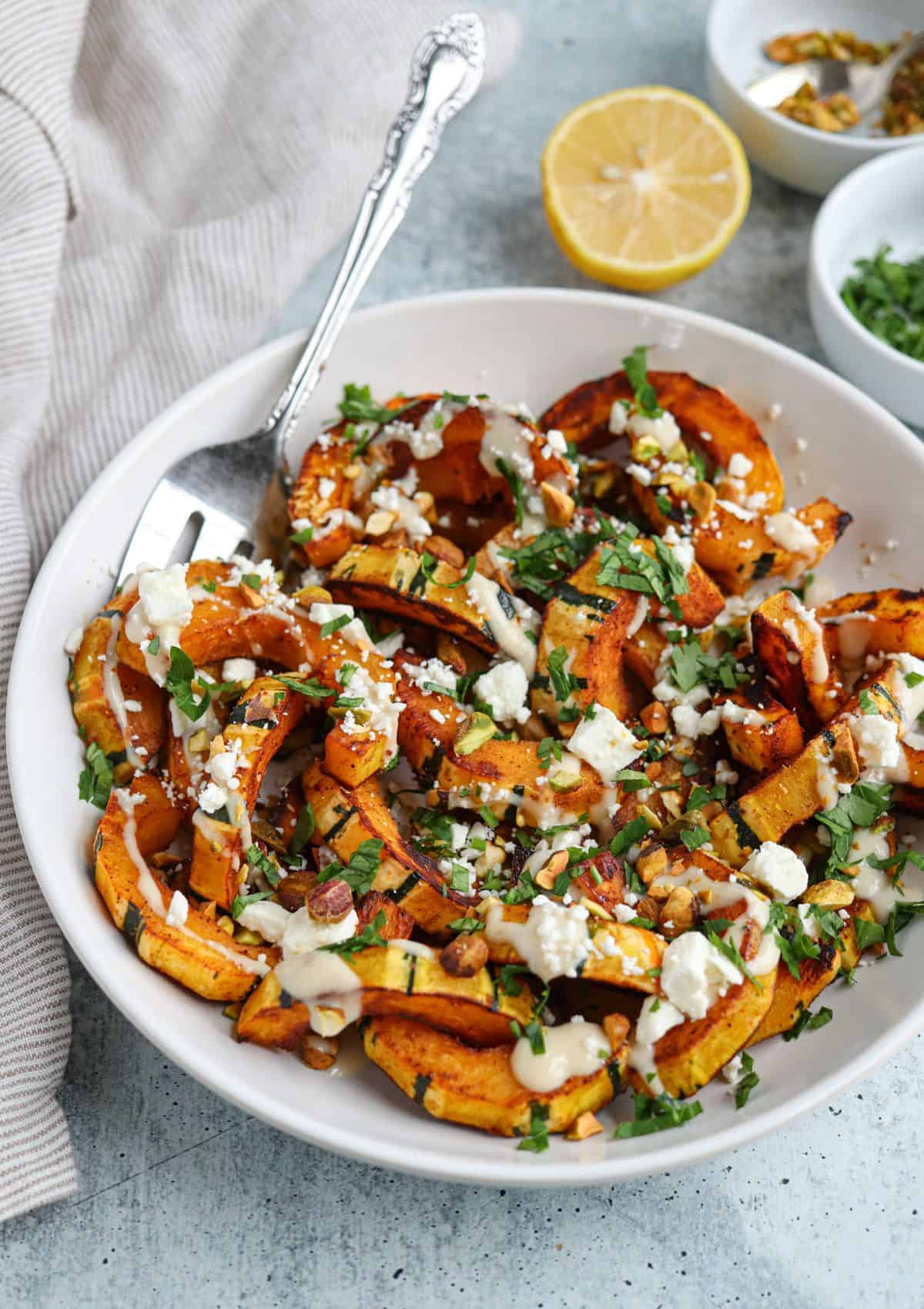 sautéed delicata squash drizzled with lemon tahini dressing and sprinkled with chopped parsley, crumbled feta cheese and pistachios in a white bowl. 