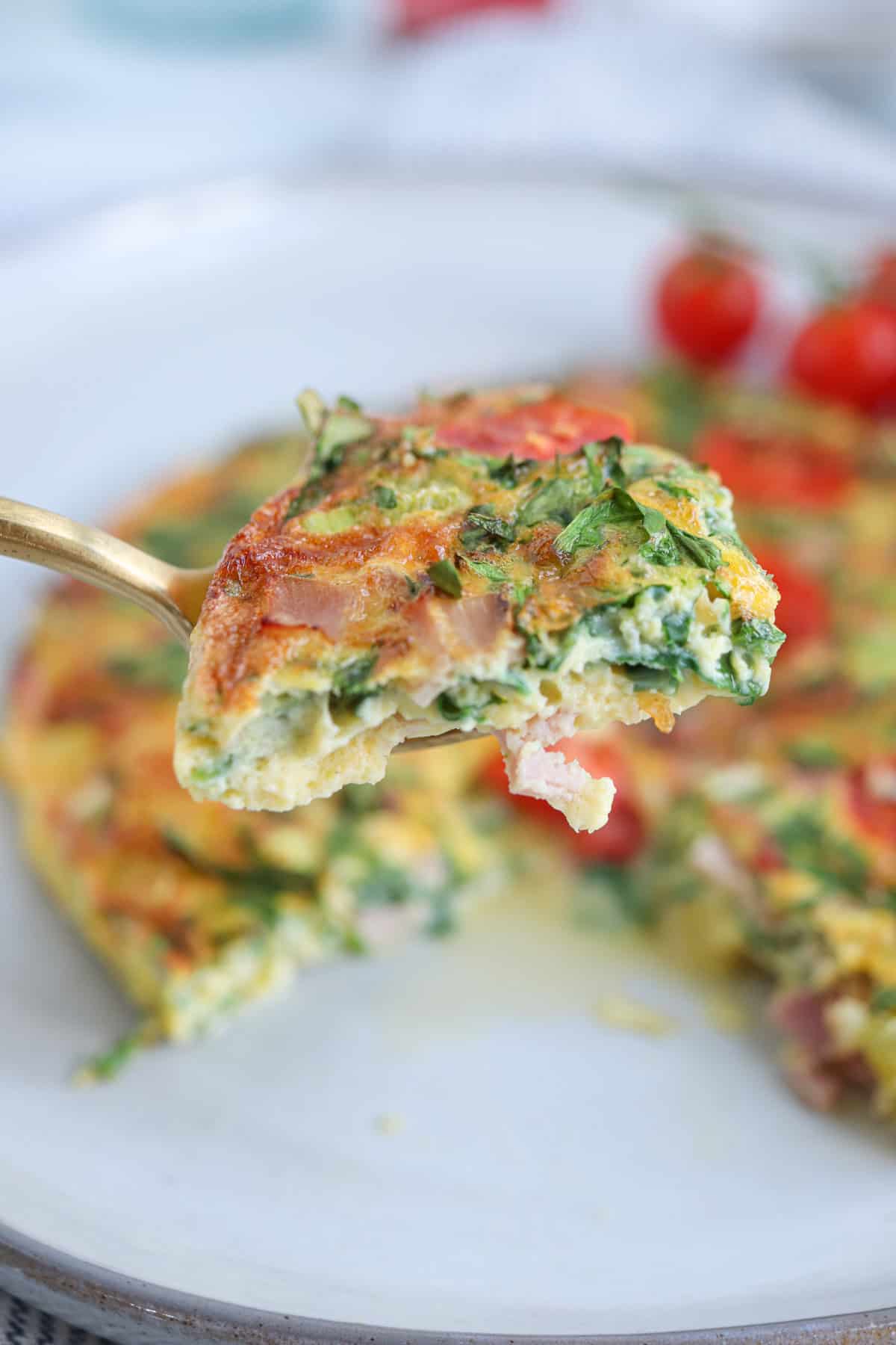 an omelette made in the air fryer with cheese, ham, spinach and cherry tomatoes on a light gray plate,  piece lifted on a fork.