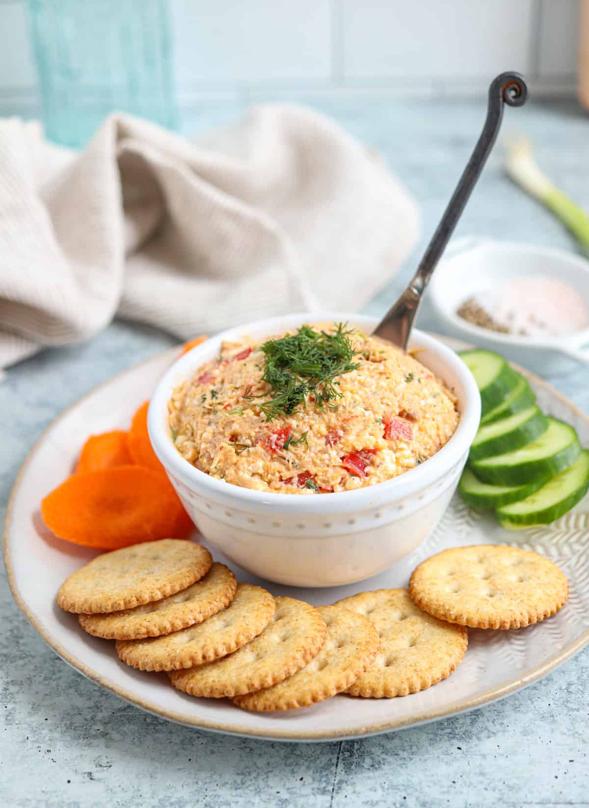 tuna spread in a white bowl on a plate surrounded by vegetables and crackers. 