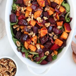 A white ceramic dish showcases a vibrant salad with roasted sweet potatoes, beets, walnuts, and mixed greens reminiscent of a falafel platter. Nearby, a small bowl of walnuts rests on the light surface.