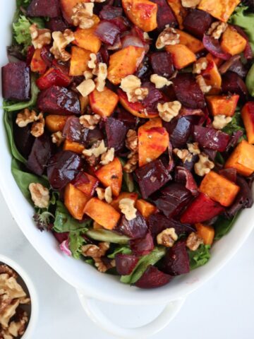 A white ceramic dish showcases a vibrant salad with roasted sweet potatoes, beets, walnuts, and mixed greens reminiscent of a falafel platter. Nearby, a small bowl of walnuts rests on the light surface.