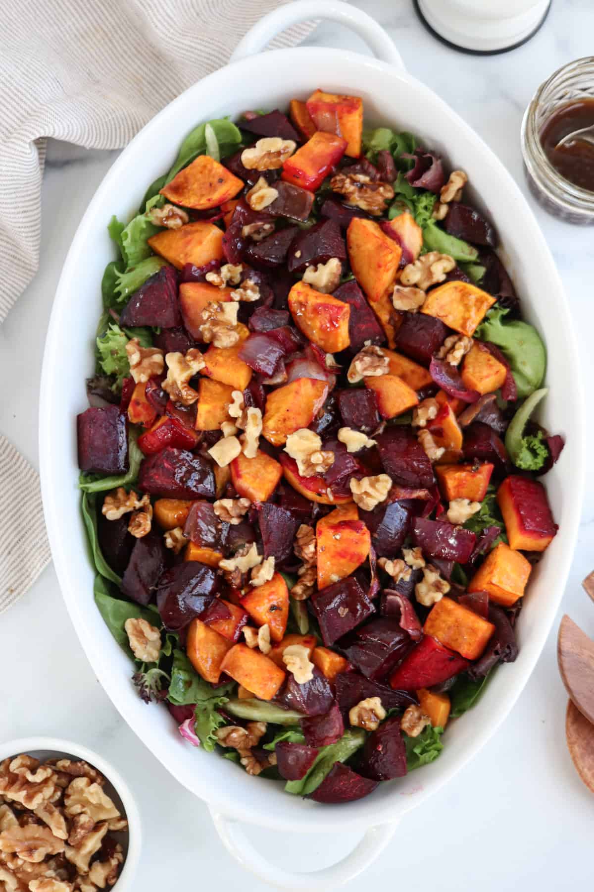 A tantalizing salad in a white dish showcases roasted sweet potatoes, beets, and walnuts over mixed greens. A jar of dressing and a small bowl of walnuts rest beside the dish on a marble surface.
