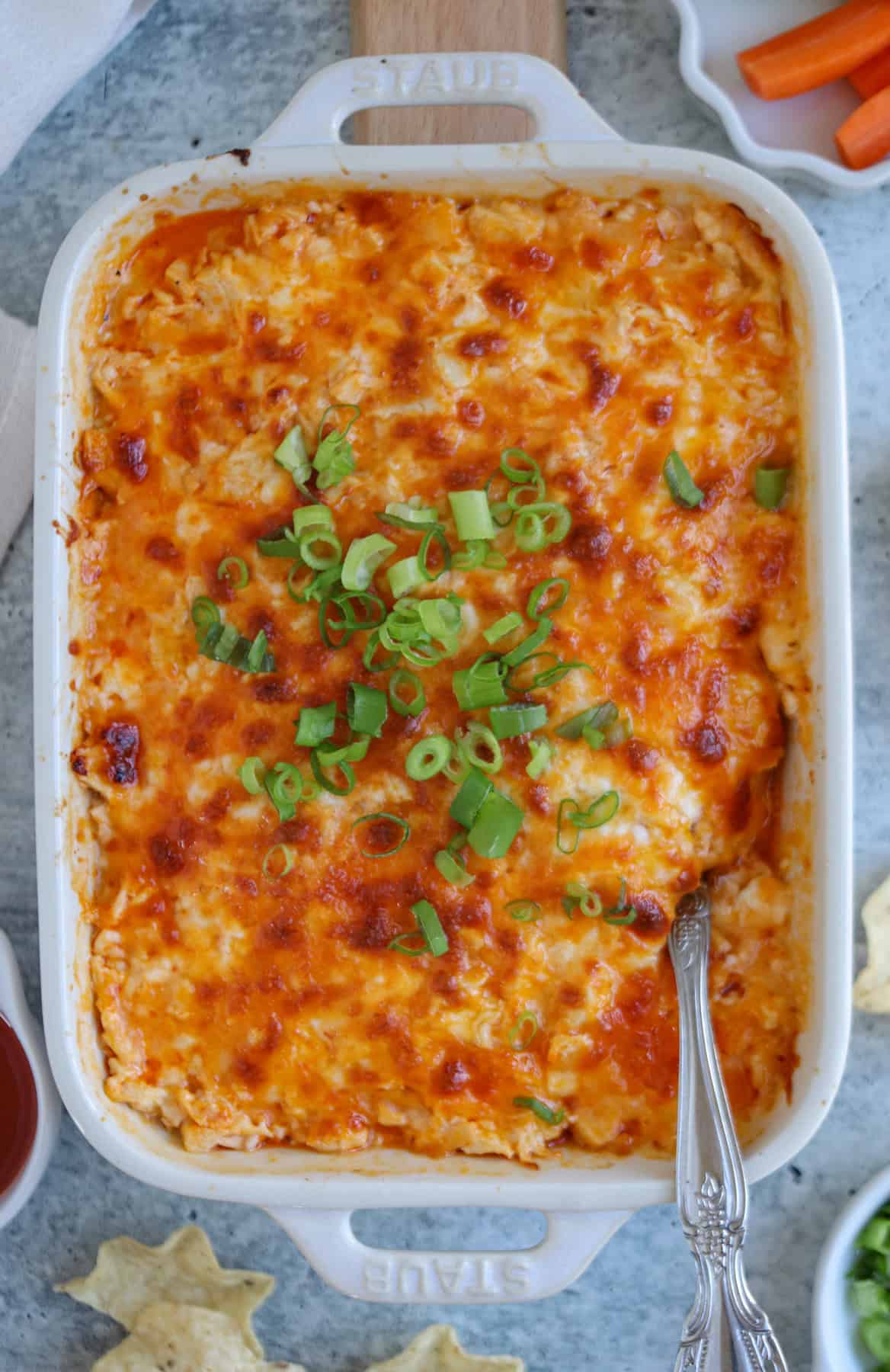 A baked casserole dish filled with cheesy buffalo chicken dip, topped with chopped green onions, sits invitingly. A serving spoon is placed in the dish. Surrounding the platter are ingredients like tortilla chips and carrot sticks on a gray surface.