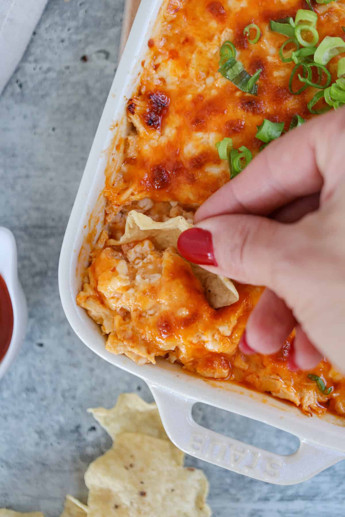 A hand with red nail polish dips a chip into a creamy, golden-brown buffalo chicken dip topped with green onions in a white baking dish. The dish sits on a gray surface with more chips nearby.