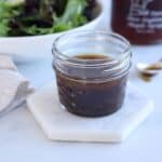A small glass jar of maple balsamic vinaigrette dressing sits on a hexagonal marble coaster. A bowl of fresh salad greens and a striped cloth napkin are partially visible in the background, along with a spoon smeared with the dressing.