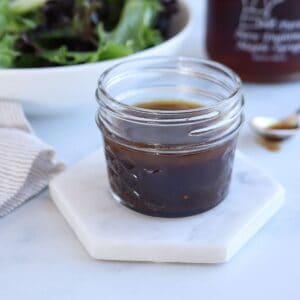 A small glass jar of maple balsamic vinaigrette dressing sits on a hexagonal marble coaster. A bowl of fresh salad greens and a striped cloth napkin are partially visible in the background, along with a spoon smeared with the dressing.