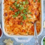 A close-up of a baked buffalo chicken dip in a rectangular dish, topped with melted cheese and chopped green onions, invites indulgence. A silver spoon rests in the dish, ready for serving. Nearby are chips and a small bowl of red sauce, reminiscent of accompaniments for a falafel platter.