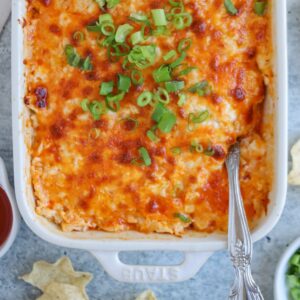 A close-up of a baked buffalo chicken dip in a rectangular dish, topped with melted cheese and chopped green onions, invites indulgence. A silver spoon rests in the dish, ready for serving. Nearby are chips and a small bowl of red sauce, reminiscent of accompaniments for a falafel platter.