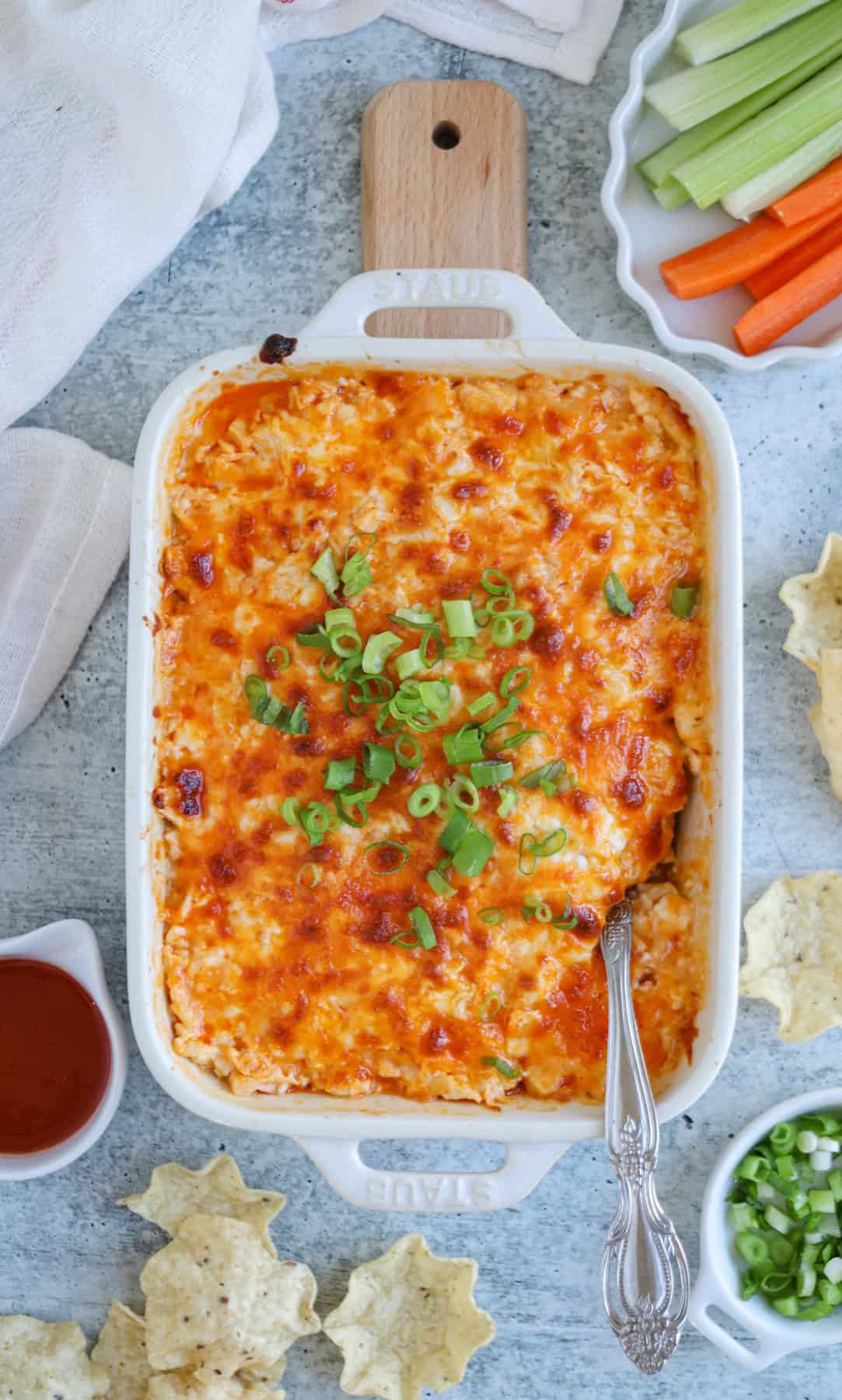 A baking dish filled with cheesy buffalo chicken dip, topped with chopped green onions, reminiscent of a vibrant falafel platter. Surrounding the dish are tortilla chips, a bowl of hot sauce, carrot and celery sticks on a plate, and a small bowl of sliced green onions.