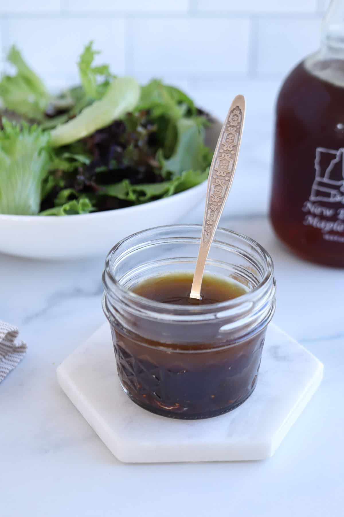 A small jar of balsamic maple vinaigrette with a silver spoon rests on a white hexagonal coaster beside a fresh falafel platter. In the background, there is a bowl of mixed greens and a bottle of Maple Syrup on the marble countertop.