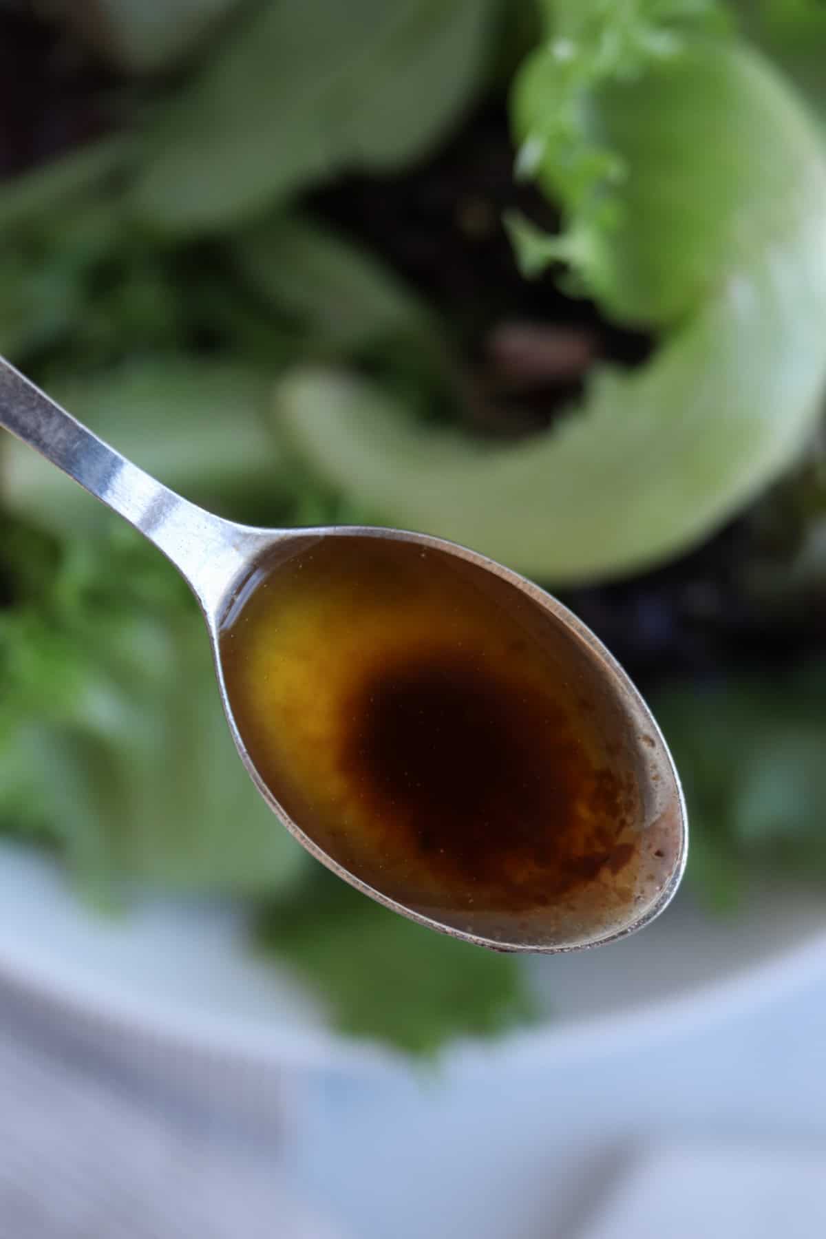 A spoon filled with dark, glossy maple balsamic vinaigrette dressing hovers above a blurred background, accompanied by fresh green lettuce leaves.