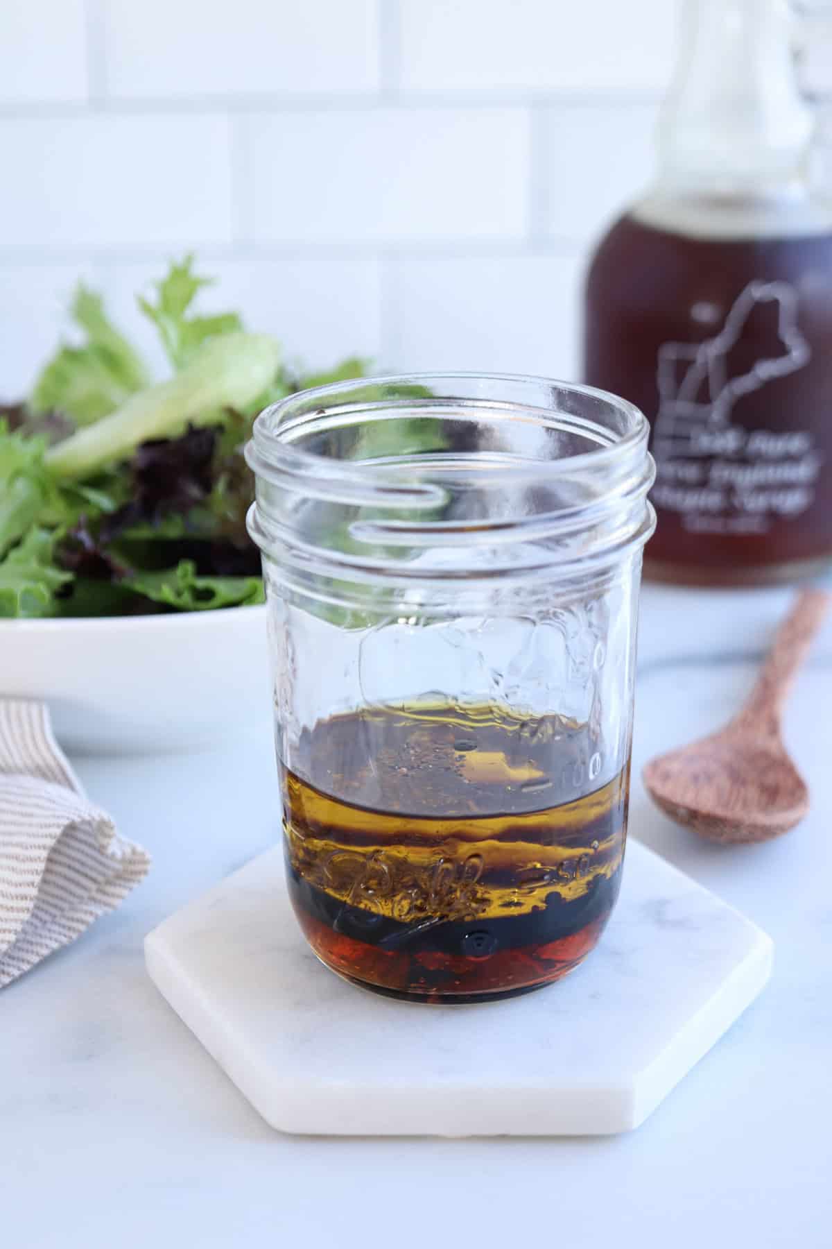 A glass jar with layers of oil and vinegar sits on a hexagonal coaster. Next to it, a bowl of mixed greens, wooden spoon, and bottle of vinaigrette in the background. The serene setup rests on a white marble countertop.