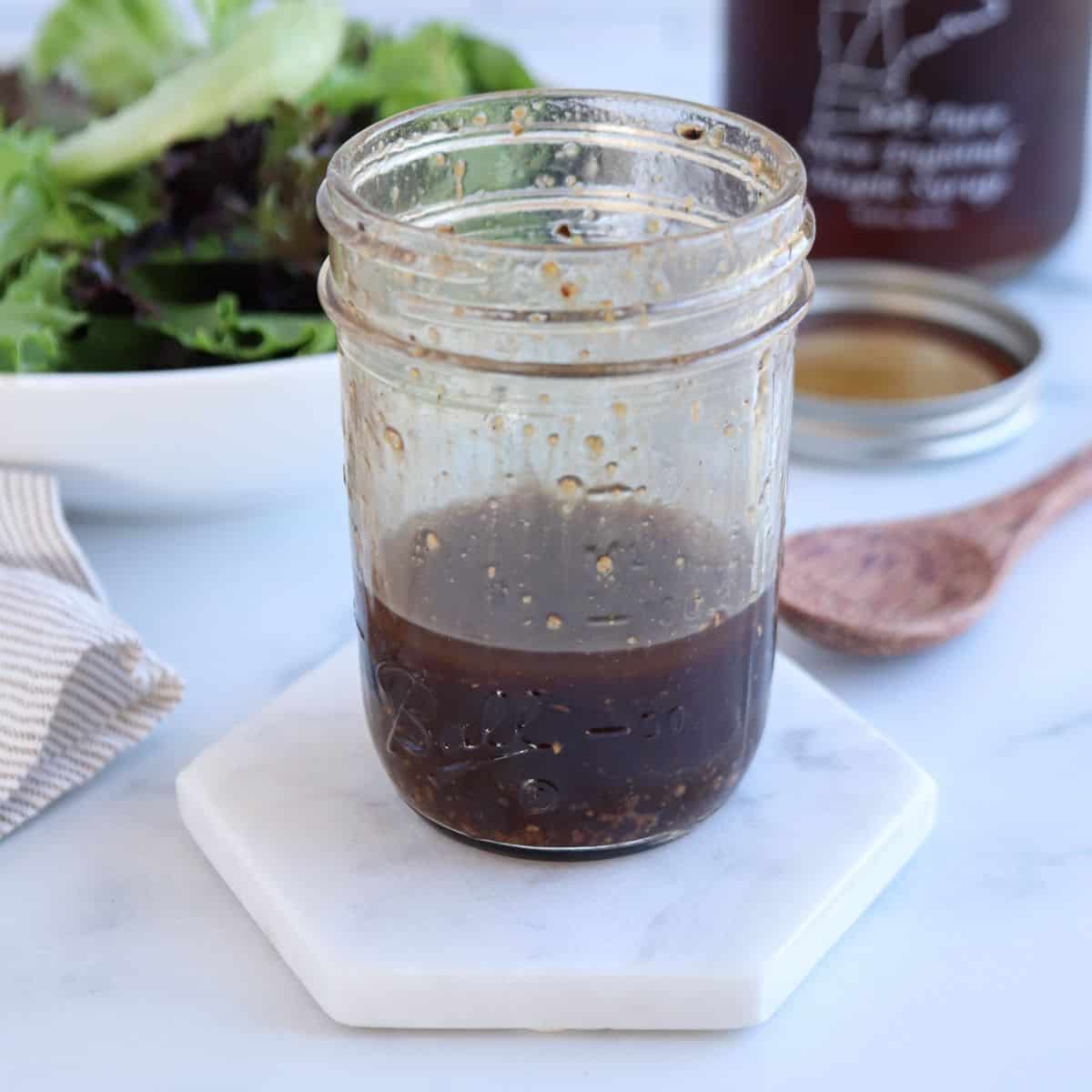 A glass jar filled with brown salad dressing sits on a white hexagonal coaster, perfect for drizzling over a falafel platter. In the background, there’s a bowl of fresh greens, a wooden spoon, and another jar with a lid. A beige striped cloth is partially visible on the left.