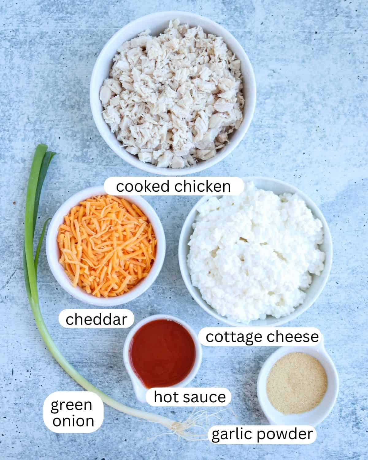 Ingredients for making buffalo chicken dip: bowls of cooked chicken, cottage cheese, and shredded cheddar alongside a small bowl of hot sauce. 