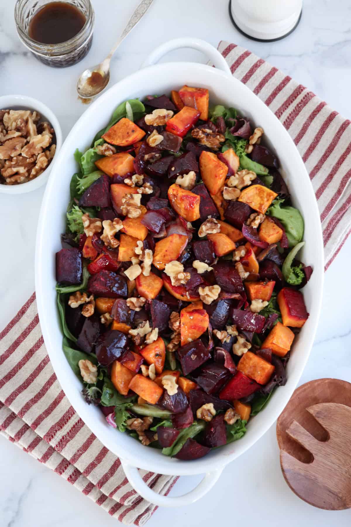 A white oval dish presents a vibrant salad of roasted beets, sweet potatoes, and walnuts over mixed greens. It rests on a red and white striped cloth, accompanied by a wooden spoon and dressing jar on the marble surface.