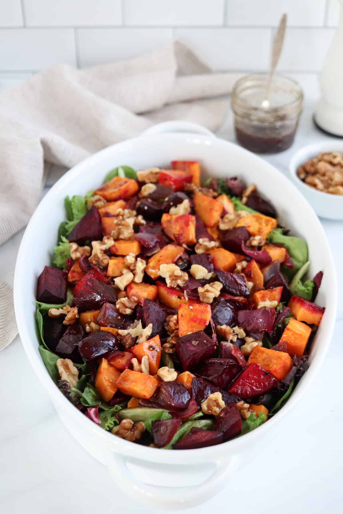 A white oval dish features a vibrant salad of roasted beets, sweet potatoes, and mixed greens, garnished with walnuts and dried cranberries. A jar of dressing and a bowl of walnuts waits in the background.